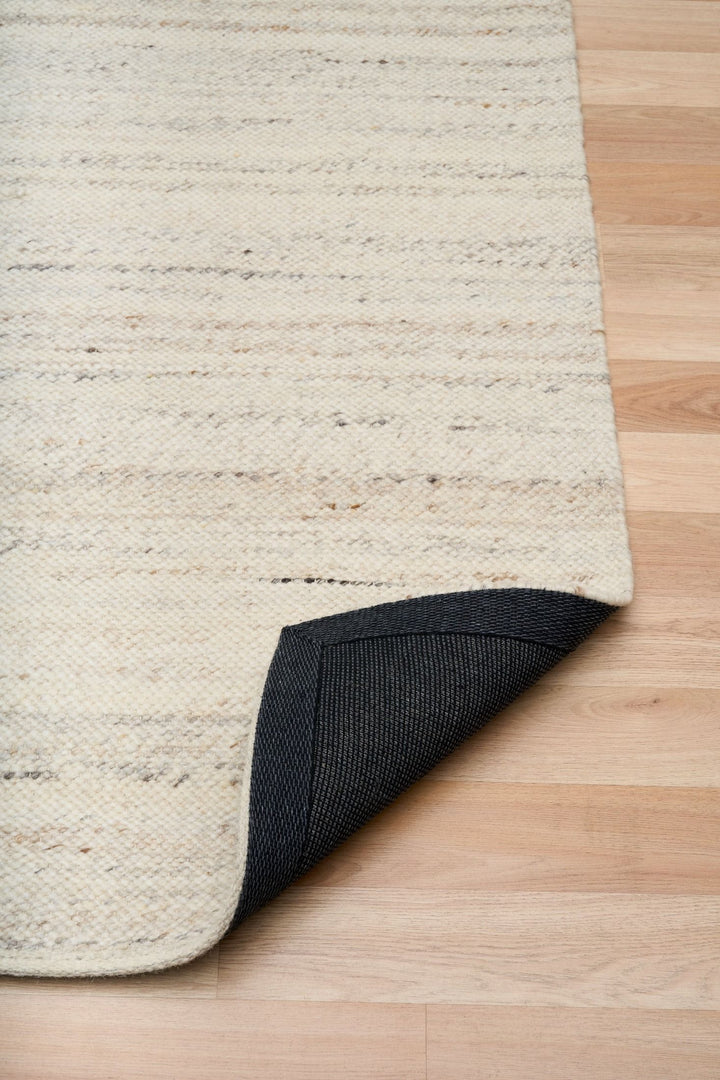 Corner of a beige woven rug lifted to show its black underside backing.
