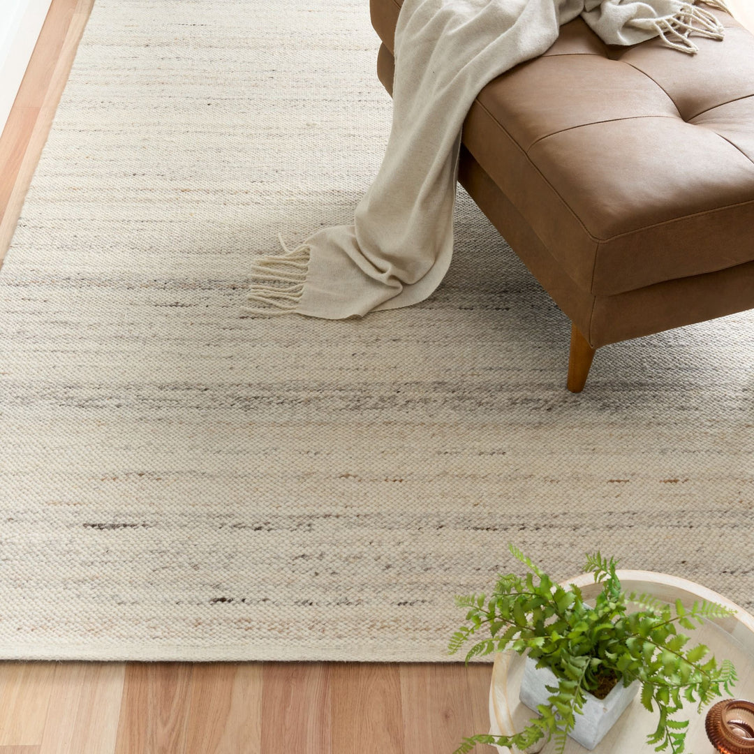 Square view of an ivory rug with a brown ottoman and green plant on a wooden floor.
