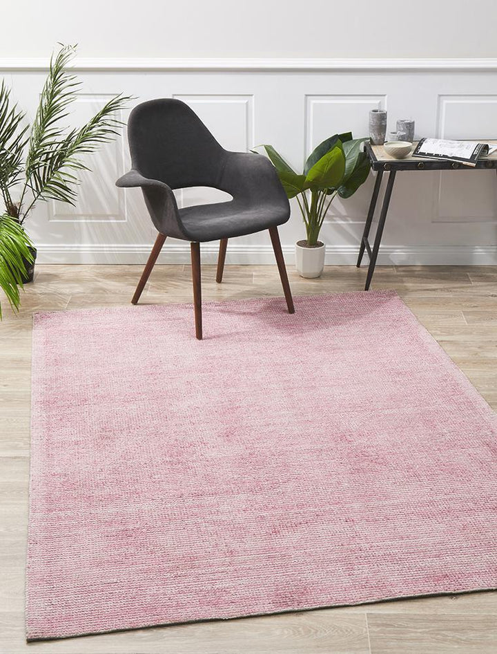 A rectangular, pink cotton and rayon rug with a modern design, featuring a black chair and wooden legs in the background.