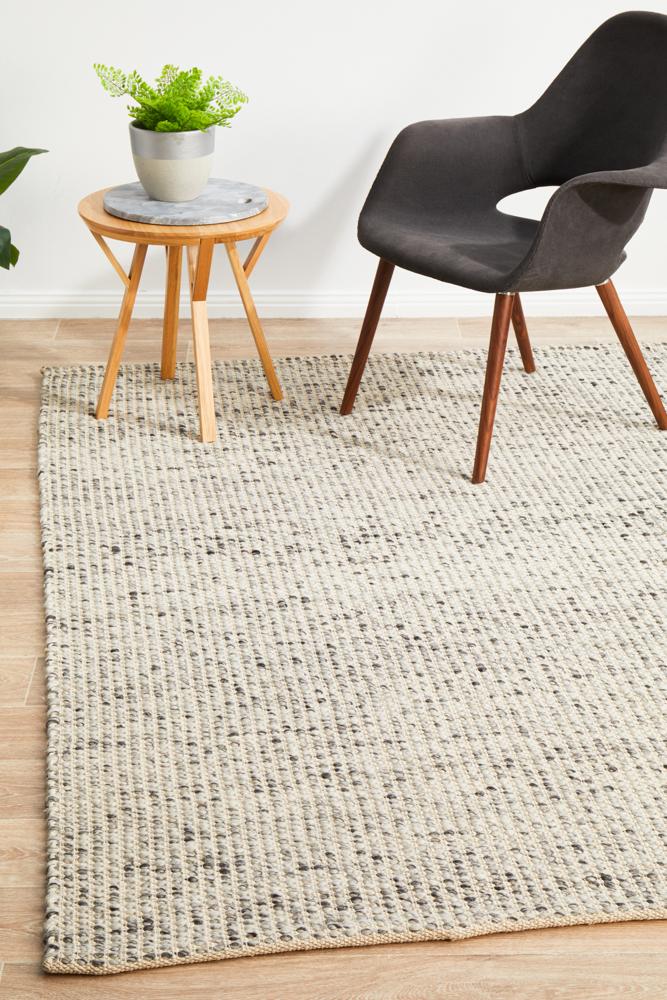 A rectangular, flatweave wool rug in gray and black, displayed on a wooden floor with a round wooden side table and a black chair in the background, indicating a minimalist interior design.