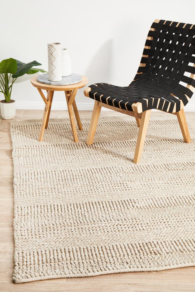 A flatweave grey rug with a Skandinavian design, displayed on a wooden floor with a black and white patterned chair and a round wooden side table in the background.