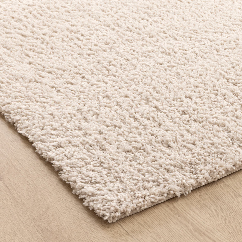 A close-up of a thick, shaggy area rug in a warm cream or off-white color, shown resting on a light wood floor.