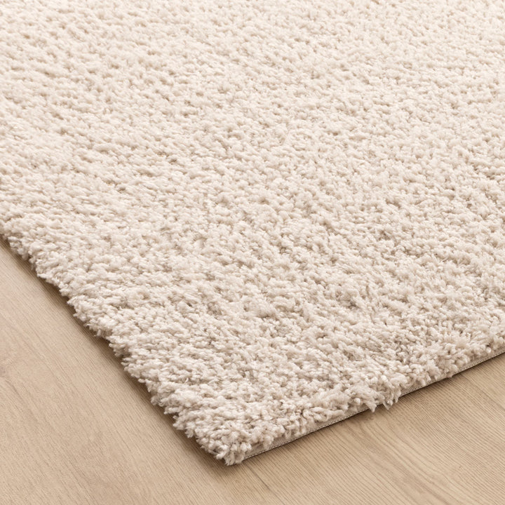 A close-up of a thick, shaggy area rug in a warm cream or off-white color, shown resting on a light wood floor.