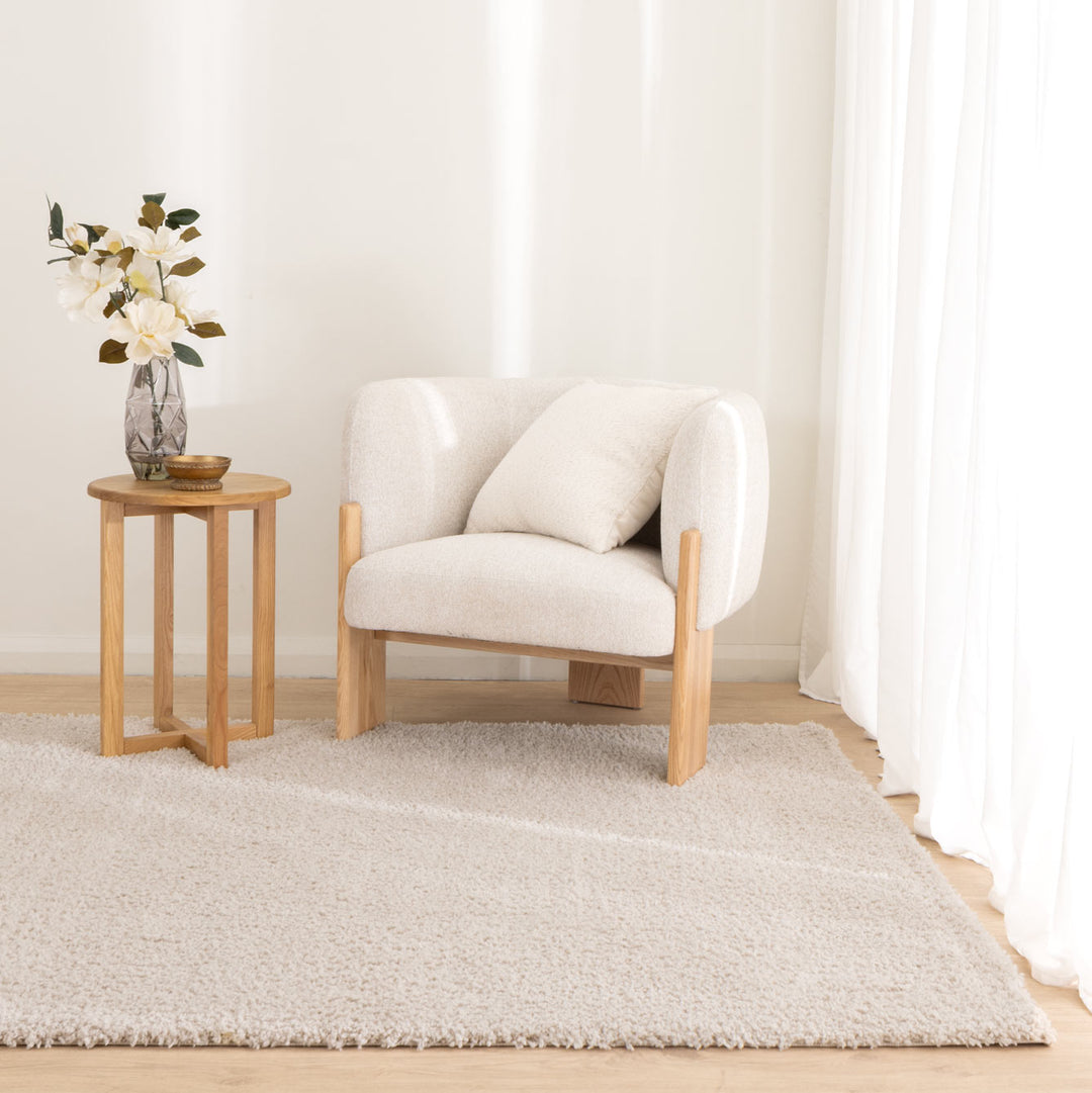 A light beige upholstered armchair with a matching cushion and natural wood frame, styled in a bright room next to a small wooden side table with a vase of flowers, all set on a shag rug.