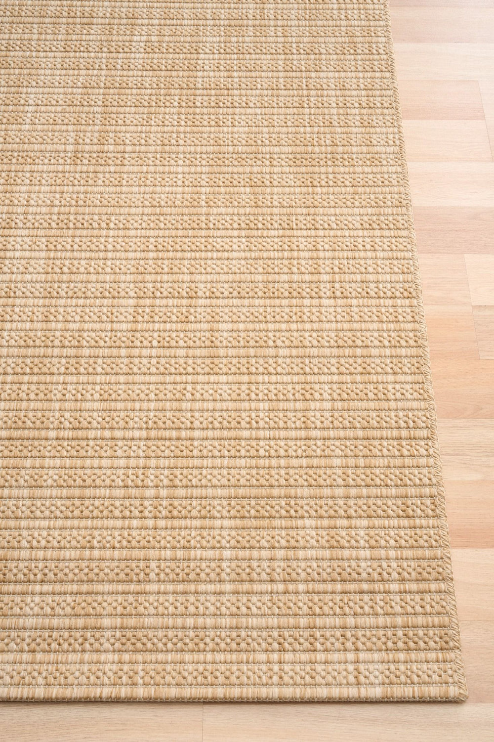 Corner view of a beige woven rug with a subtle diamond lattice pattern, serged edge detail, on light wood flooring.