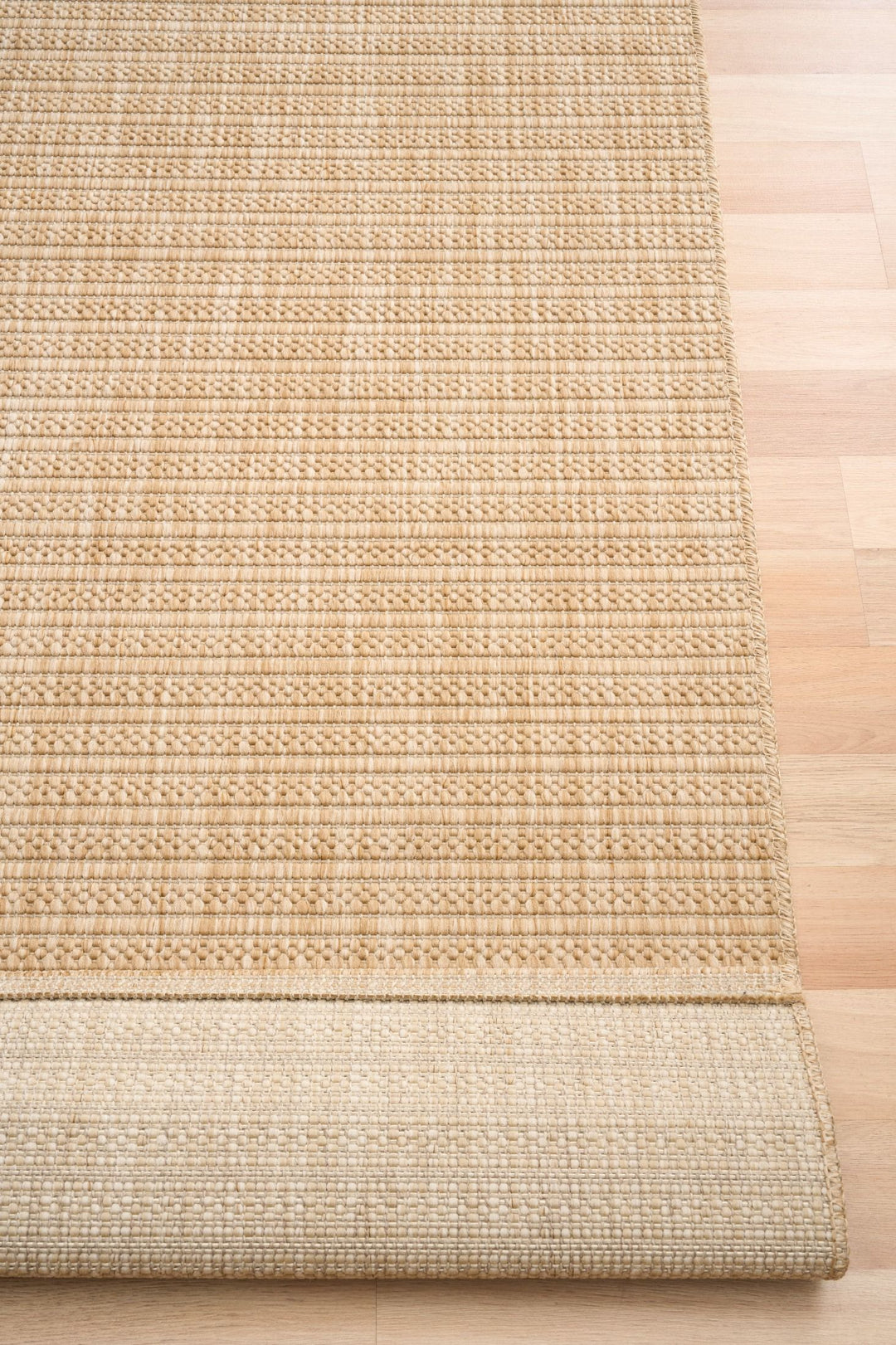 Close-up of tan woven rug with diamond lattice, serged edge, and rolled underside on wood floor.