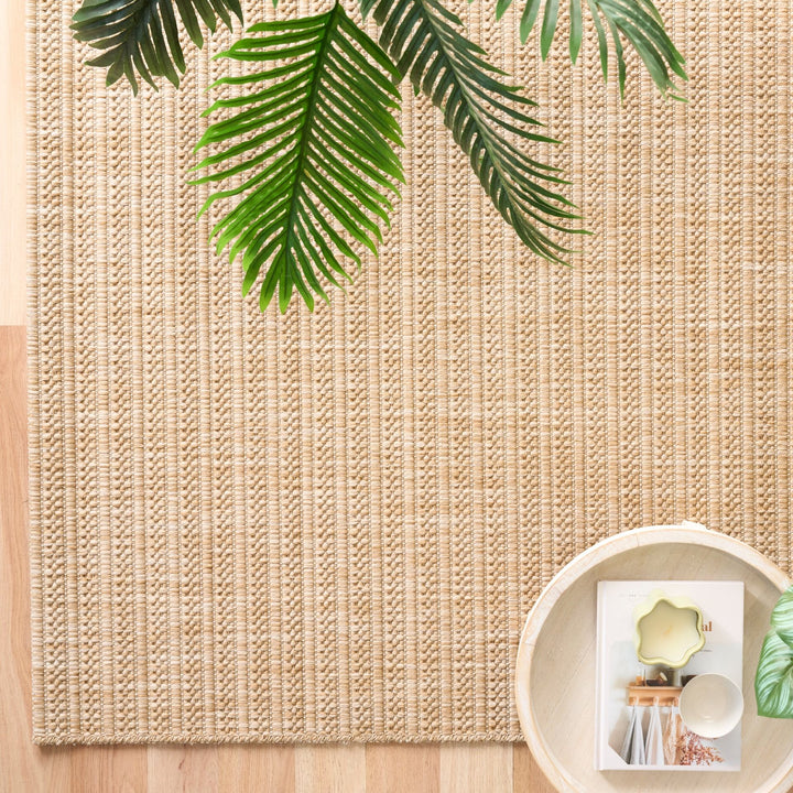 Tan flat-woven rug with ribbed stripes, styled with palm leaves and a tray on light wood flooring.