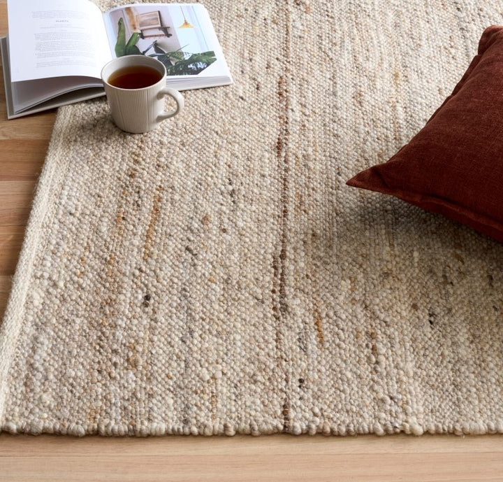Beige textured rug styled in a cozy room with a cup of tea, open book, and cushion."