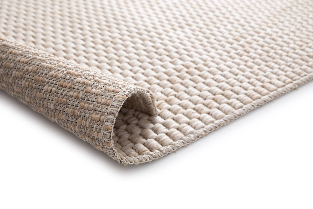 Close-up of a light beige woven rug with a rolled corner, showing basketweave underside, looped pebble texture, and stitched edge on a white background.