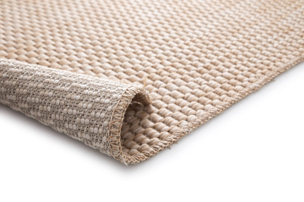 Close-up of the Barton indoor/outdoor rug in Sand (41503-070), rolled corner showing woven backing, chunky basketweave texture, and serged edge on a white background.
