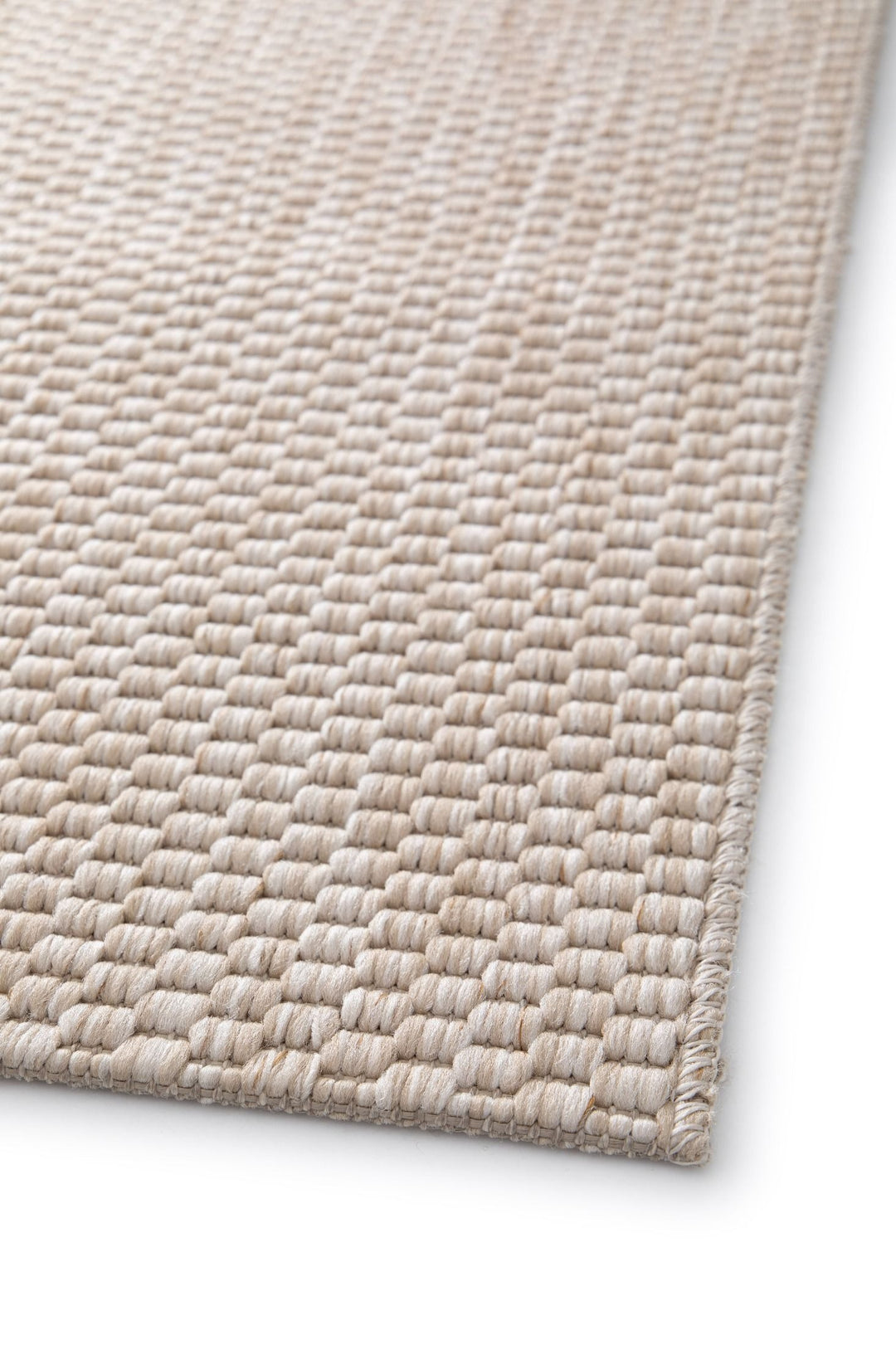 Close-up angled view of a light beige pebble-weave area rug corner with overlocked edge, showing the thick looped woven texture on a white background.