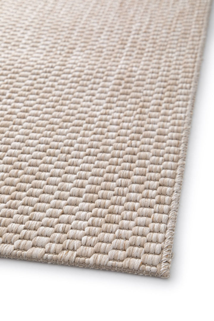Close-up angled view of a light beige pebble-weave area rug corner with overlocked edge, showing the thick looped woven texture on a white background.