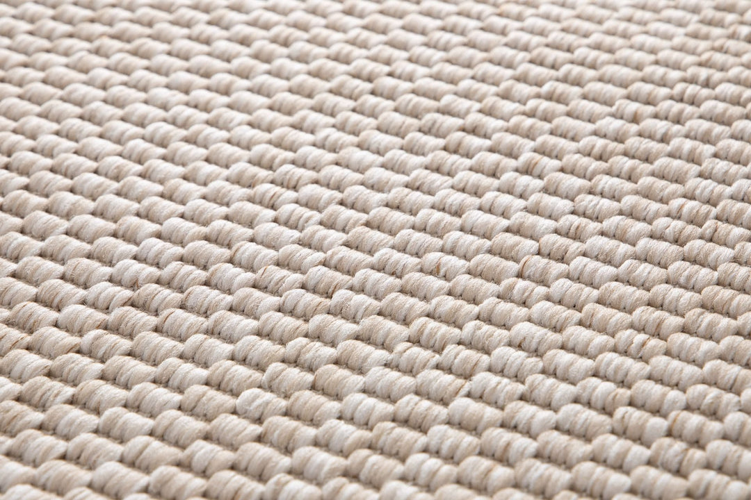Macro close-up of a light beige pebble-weave area rug, showing thick looped yarn rows and textured neutral cream–tan fibers.