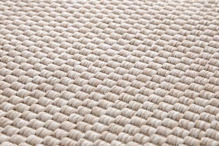 Macro close-up of a light beige pebble-weave area rug, showing thick looped yarn rows and textured neutral cream–tan fibers.