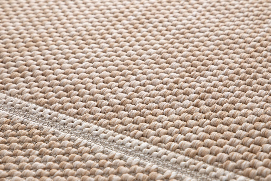 Macro close-up of a beige woven area rug showing raised looped basket-weave texture with a stitched border seam running diagonally; sand and ivory tones (Barton 41505/70).