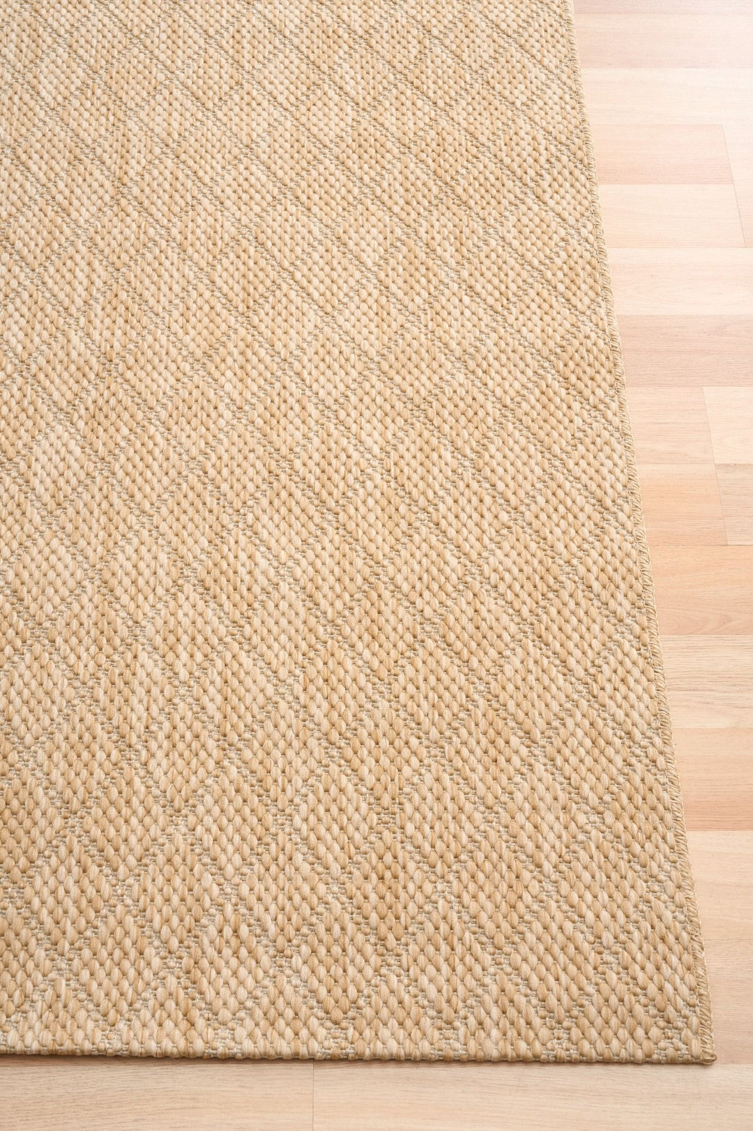 Corner view of beige woven rug with diamond lattice pattern and serged edge on light wood floor.