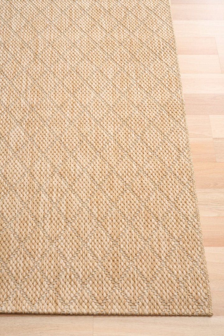 Corner view of beige woven rug with diamond lattice pattern and serged edge on light wood floor.