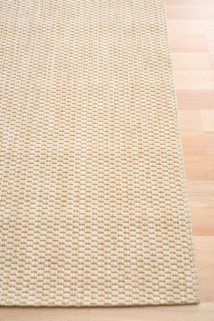 Corner detail of a light beige/ivory woven area rug with chunky pebble texture and serged edge, shown on light wood flooring.