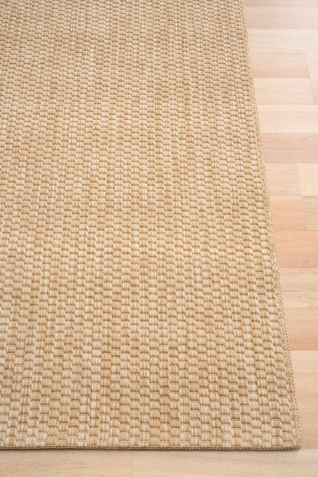 Corner detail of the Barton indoor/outdoor rug in Sand (41503-70) on a light wood floor, showing the thick basketweave texture and bound edge.