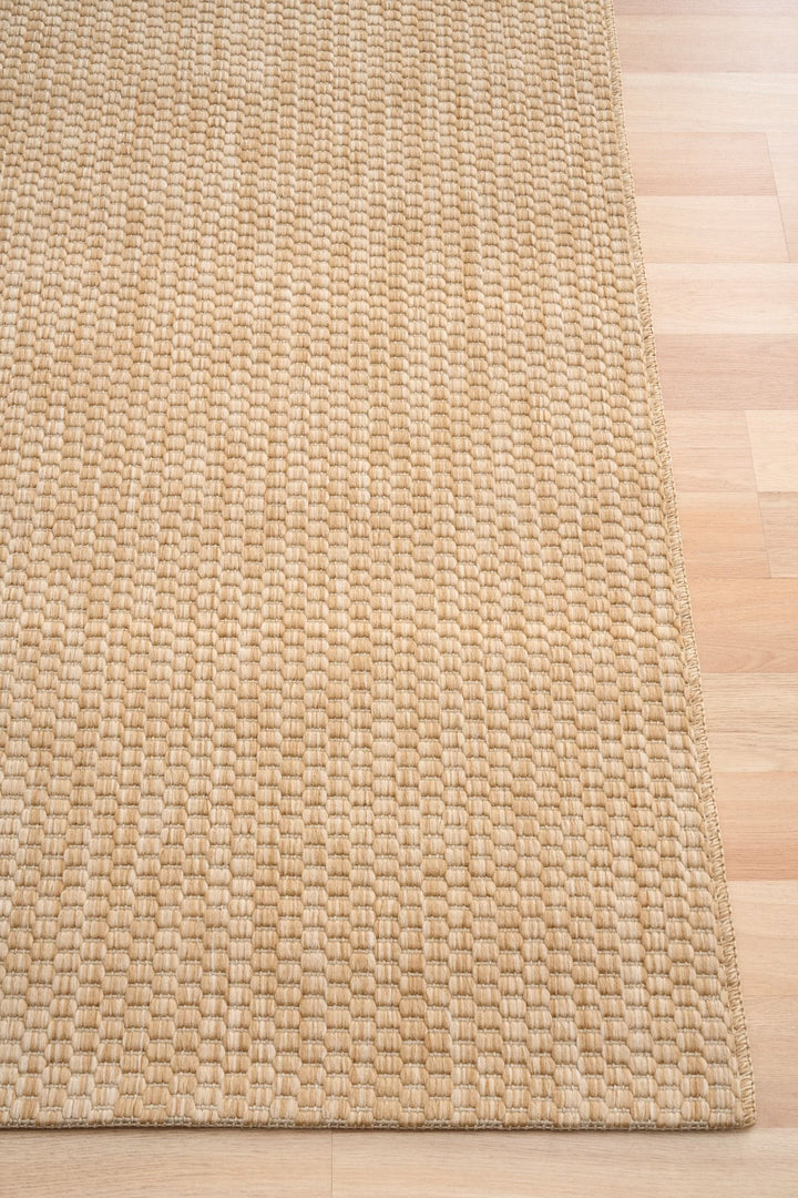Corner detail of the Barton indoor/outdoor rug in Sand (41503-70) on a light wood floor, showing the thick basketweave texture and bound edge.