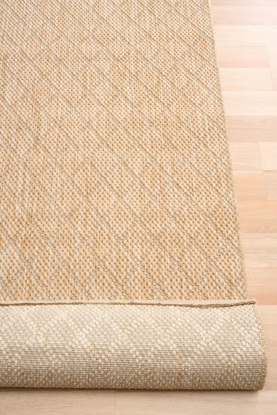 Close-up of tan woven rug with diamond lattice, serged edge, and rolled underside on wood floor.