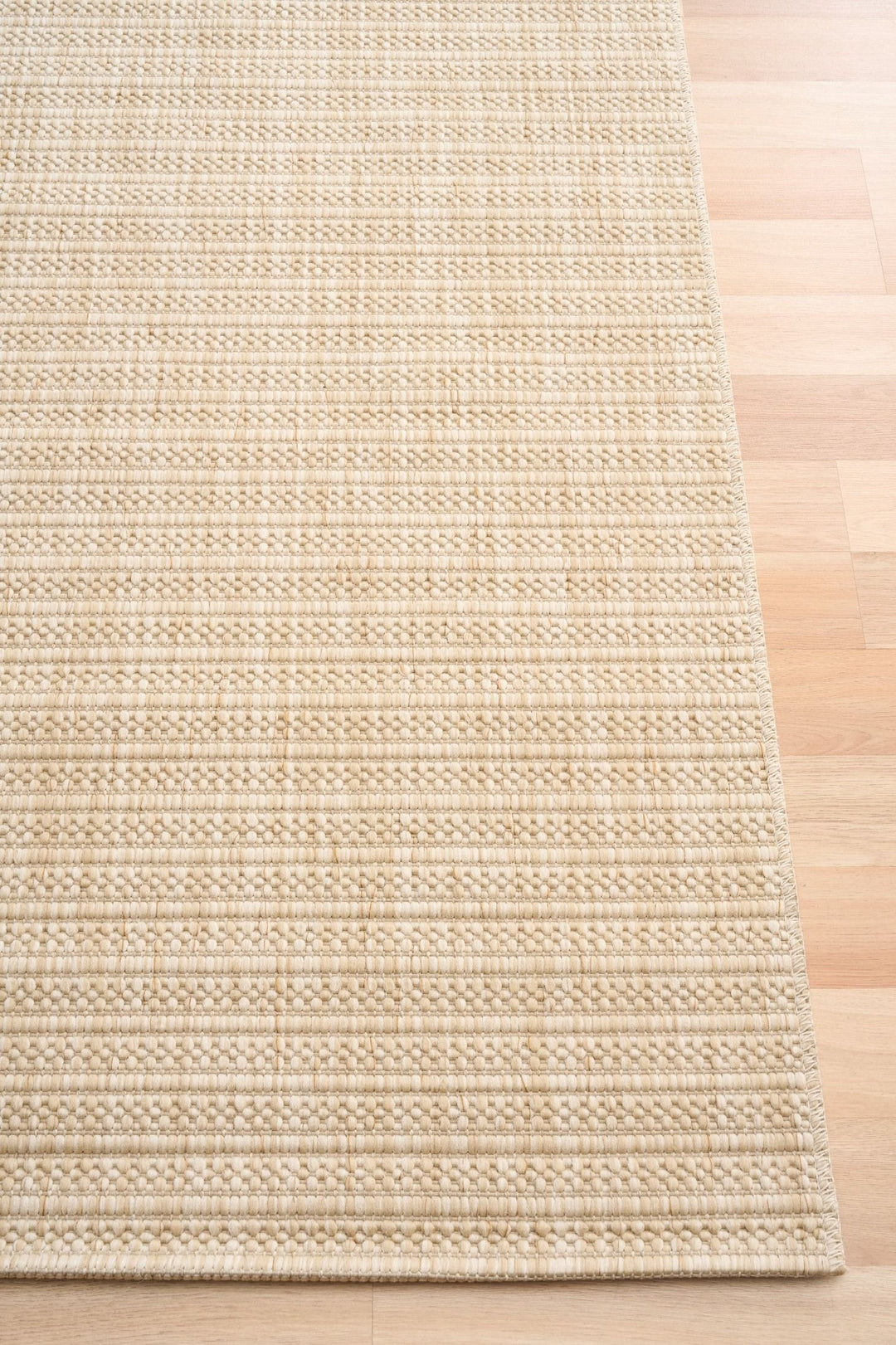 Corner view of a beige woven rug with a subtle diamond lattice pattern, serged edge detail, on light wood flooring.