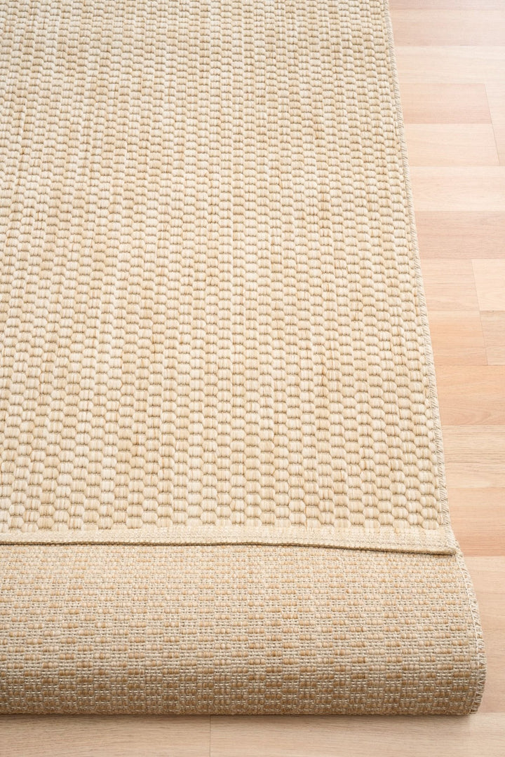Rolled edge of a light beige/ivory woven area rug with chunky basket-weave texture and serged border, showing the underside on light wood flooring.