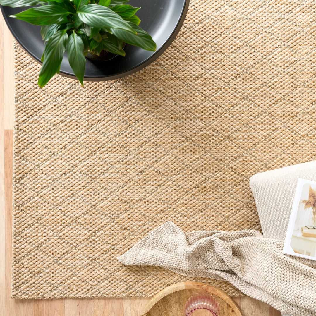 Top-down view of tan woven rug with diamond lattice, styled with planter, throw, and tray on wood floor.