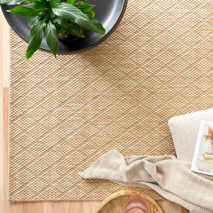 Top-down view of tan woven rug with diamond lattice, styled with planter, throw, and tray on wood floor.