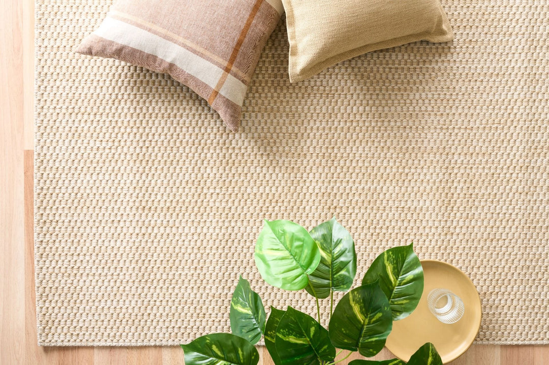 Top-down view of a light beige woven area rug with pebble texture, bordered by hardwood floor, styled with two neutral throw pillows, a green houseplant, and a gold tray with a small glass candle.