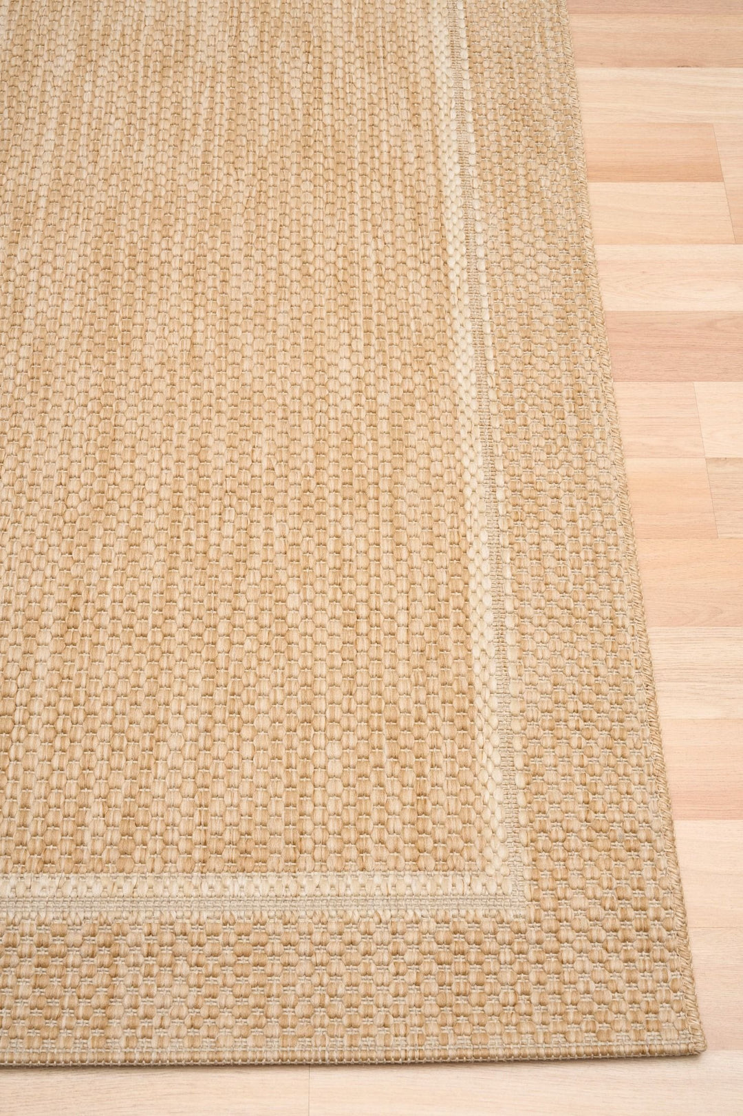 Close-up of a beige/tan woven area rug corner on a light wood floor, showing the textured loop weave and lighter double border detail.