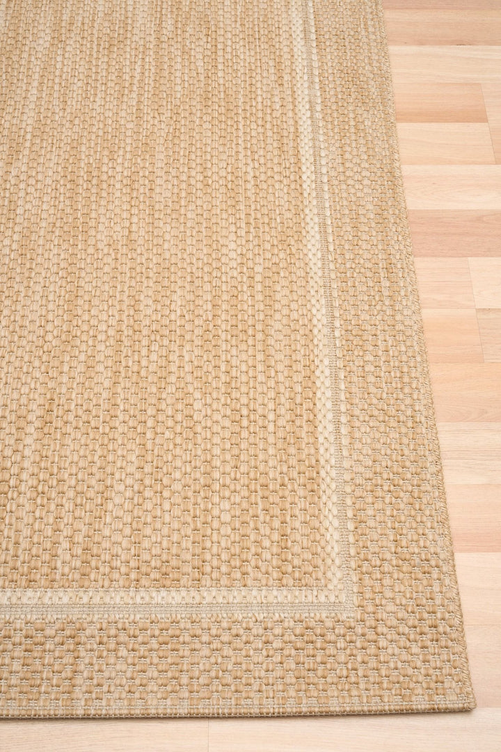 Close-up of a beige/tan woven area rug corner on a light wood floor, showing the textured loop weave and lighter double border detail.