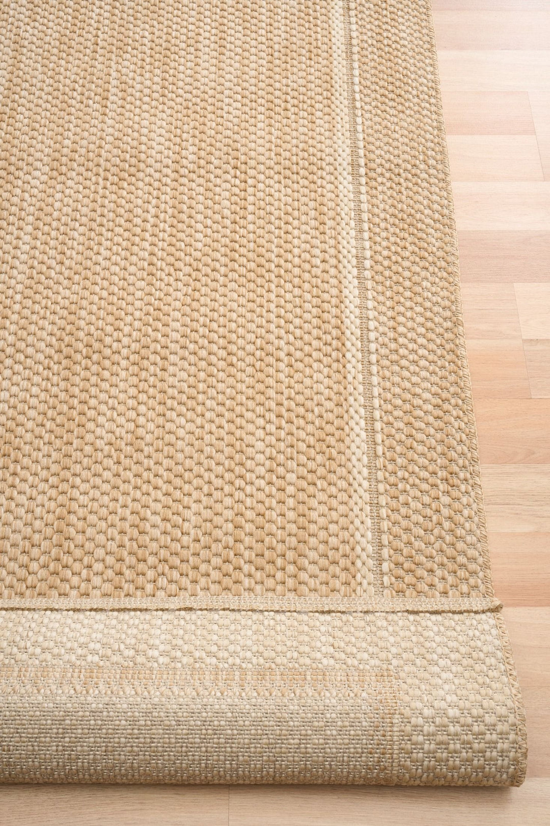 Rolled edge of a beige woven area rug on a light wood floor, showing the textured loop weave, lighter double border, and the underside pattern at the corner.