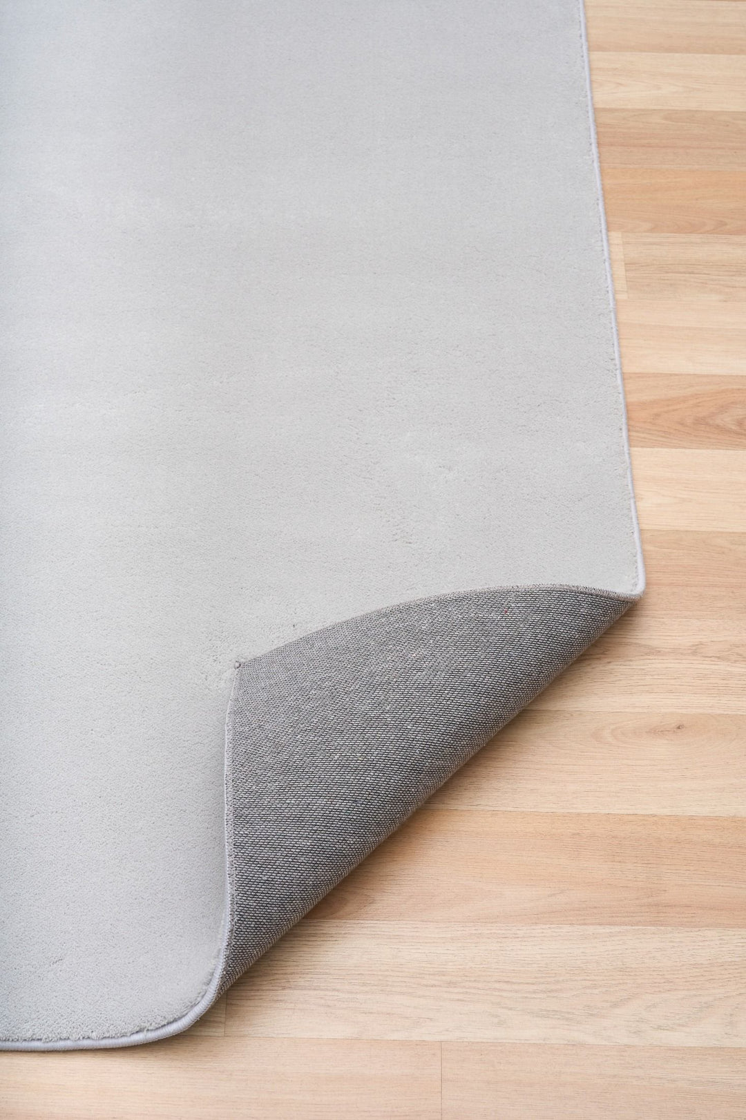 Light grey rug corner lifted to reveal its textured underside on a wooden floor.