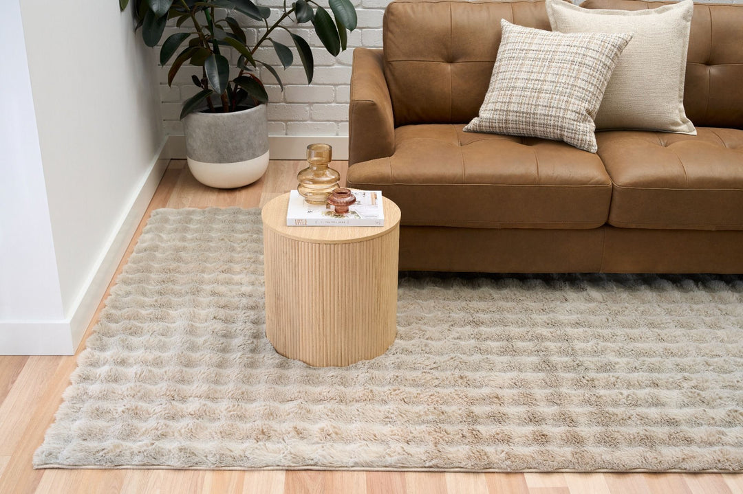 Light beige textured rug in living room with brown leather sofa, pillows, and round wooden side table.