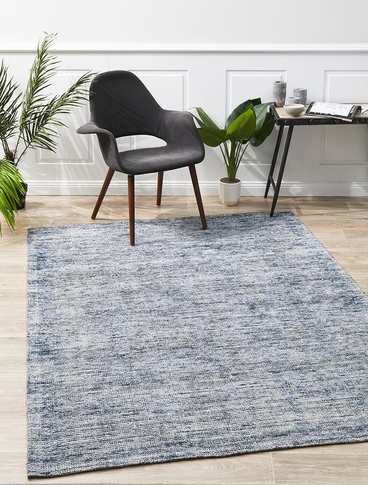 An indigo cotton and rayon rug with a modern design, displayed in a room with a black chair and green plants in the background.