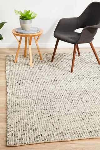 A rectangular, flatweave wool rug in gray and black, displayed on a wooden floor with a round wooden side table and a black chair in the background, indicating a minimalist interior design.